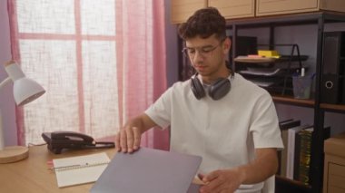 Ofisteki genç İspanyol adam dizüstünü kapatıp kulaklık takıyor, müzik dinlemeye hazırlanıyor ve kapalı bir iş yerinde dinleniyor..