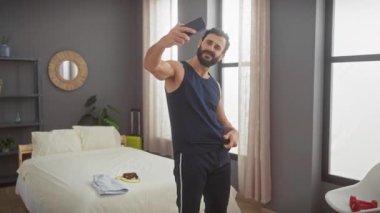 Sakallı yakışıklı bir adam şık bir yatak odasında karın kaslarını göstererek selfie çekiyor..