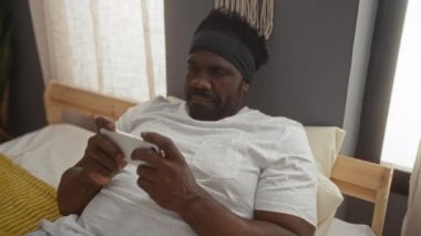 Sakallı, Afro-Amerikalı genç bir adam akıllı telefonunu kullanarak yatakta uzanıyor, rahat bir yatak odasında çeşitli duygular sergiliyor..