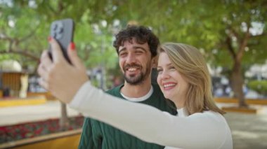 Neşeli bir adam ve kadın, ağaçlar ve banklarla çevrili bir parkta selfie çekiyorlar..