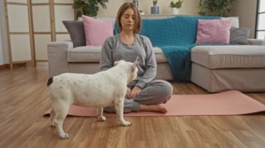 Oturma odasında evcil köpeğiyle yoga yapan genç bir kadın. Etrafı modern mobilya ve bitkilerle çevrili ahşap bir zeminde.