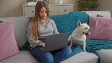 Genç sarışın bir kadın, rahat bir oturma odasında köpeğinin yanındaki koltukta otururken dizüstü bilgisayar kullanıyor..