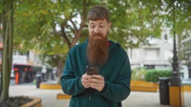 Sakallı bir adam parkta akıllı bir telefonla heyecanını dile getiriyor.