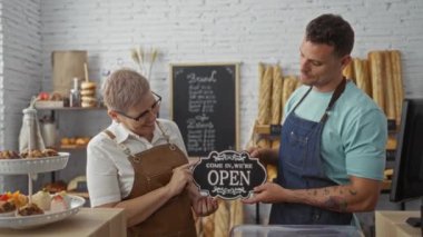 Kadın ve erkek fırıncılar fırının içinde açık tabela tutuyorlar ve arka planda ekmek ve hamur işleriyle birlikte iş için hazır olduklarını gösteriyorlar.