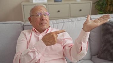 Gri saçlı yaşlı bir adam oturma odasındaki bir kanepede pembe gömlekli, iç mekan bitkileri ve arka planda bir dolap olan bir koltukta otururken gülümsüyor..