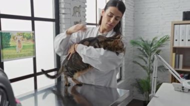 Modern bir veteriner kliniğinde kedisini tutan genç bir İspanyol kadın, arka planda tıbbi ekipmanlar ve hayvanlarla ilgili görüntülerle birlikte kapalı bir veteriner ortamını sergiliyor..