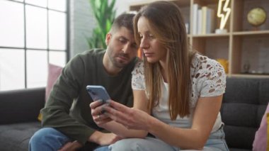 Endişeli bir çift rahat oturma odalarındaki akıllı telefonu inceliyor. İlişkiyi, teknolojiyi ve ev hayatını temsil ediyor..