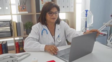 Orta yaşlı, İspanyol, iyi organize olmuş bir klinikte dizüstü bilgisayarla çalışan bir kadın doktor. Arkaplanda tıbbi ekipman ve raflar var..