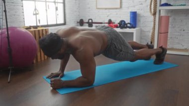 Parlak bir spor salonunda, mavi bir paspasın üzerinde şınav çeken yakışıklı bir genç..