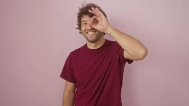 Tişörtlü, pembe izole edilmiş arka planda duran neşeli, genç İspanyol bir adam. Mutlu adam sırıtıyor, parmaklarıyla 