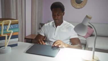 Evdeki oturma odasında dizüstü bilgisayarda çalışan genç bir adam, masa lambası ile aydınlatılmış, sehpasında bir yığın kitap ve modern iç dekorla.