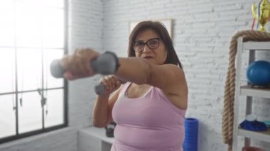 Spor salonunda dambıllarla çalışan, gözlüklü ve pembe kolsuz tişörtlü olgun bir kadın. Parlak, kapalı bir spor salonundaki egzersiz rutinine odaklanmış..