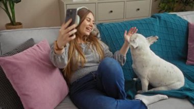 Genç, sarışın bir kadın oturma odasında kulaklık takıp, beyaz köpek köpeğiyle kanepede otururken telefonuyla video görüşmesi yapıyor..