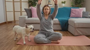 Genç sarışın kadın minderde köpek köpeğiyle yoga yapıyor. Oturma odasında sakin bir ortam sergiliyor..