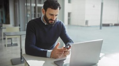 Akıllı telefon ve laptop kullanan olgun sakallı adam modern ofiste düşünceli ve stresli görünüyor..