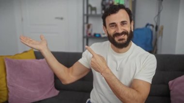 Yakışıklı orta yaşlı bir adam, şaşırmış ve gülümsüyor, kameraya kendinden emin bir şekilde bakıyor, evde beyaz bir tişört giyerken tek elini gösteriyor..