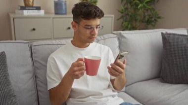 Gözlüklü genç bir İspanyol, telefonuna bakarken kahve içer. Modern bir oturma odasında, bitki ve dekorasyonla birlikte bir koltukta oturur..