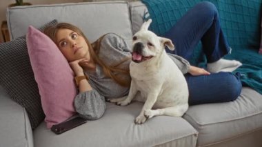 Oturma odasındaki kanepede köpeğiyle dinlenen genç bir kadın, evde akıllı telefon kullanıyor..