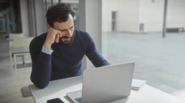 Olgun sakallı bir adam modern bir ofiste kendini yorgun ve stresli hissediyor. Masasında dizüstü bilgisayarı ve akıllı telefonu var..