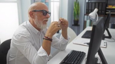 Gözlüklü orta yaşlı bir adam çift monitörlü bir ofiste çalışıyor, görevlerine odaklanıyor ve elinde bir kalem tutuyor.