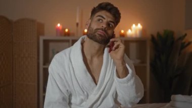 Kaplıca takımındaki yakışıklı genç adam, beyaz bir bornoz giyip telefonda konuşuyor. Arka planda mumlar var. Rahatlatıcı bir ortam için.