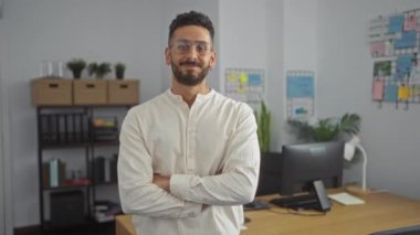 Modern bir ofis ortamında, açık renk gömlek ve gözlüklü, dekoratif ofis malzemeleriyle çevrili, kolları katlanmış bir masanın önünde duran, kendine güvenen genç İspanyol bir adam..