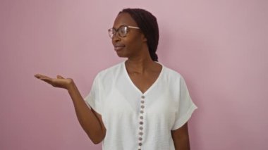 Genç Afro-Amerikalı kadın, gözlüklü, neşeli bir gülümseme takınıyor ve pembe arka planı işaret ediyor.