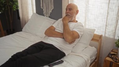 Beyaz saçlı, sakallı, rahat bir yatak odasında yatakta dinlenen beyaz tişörtlü ve siyah pantolonlu bir adam..
