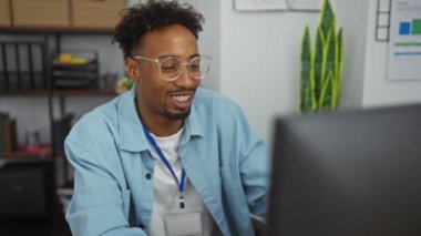 Ofiste çalışan, gülümseyen ve bilgisayarına odaklanmış, rafları ve arka planda bitkileri olan Afro-Amerikan genç bir adam..