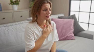 Sarışın ve endişeli bir yüzü olan güzel bir kadın oturma odasında bir kanepede oturuyor elini göğsüne bastırıyor ve bir ev ortamında tıbbi bir acil durumu vurguluyor..