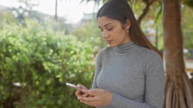Genç, mutlu İspanyol bir kadın telefonunu açık havada, yemyeşil bir park ortamında kullanır, neşe ve memnuniyeti ifade eder..
