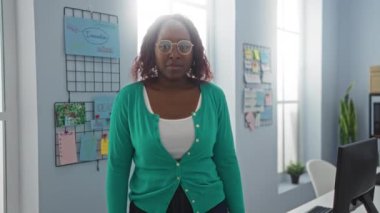 Genç, Afrikalı, Amerikalı, kadın, gözlüklü, bir ofiste, kendine güvenen, çapraz kollu, aydınlık bir odada, iş yerlerini gösteren, elementleri yerleştiren.