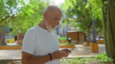 Sunny Parkta yeşil ağaçlı akıllı telefon kullanan kıdemli bir adam.