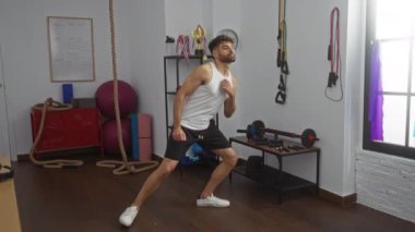 Spor salonunda spor aletleriyle egzersiz yapan genç bir adam, kolsuz tişört ve şort giyiyor, halatlar, ağırlıklar ve spor aksesuarlarıyla çevrili..