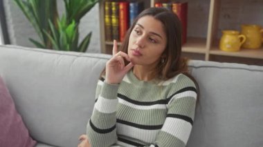 Düşünceli, genç bir İspanyol kadın, evinde bir koltukta oturuyor, yüzünde ciddi bir ifade, çenesinde bir el, kafa karıştırıcı bir soruyu düşünüyor.