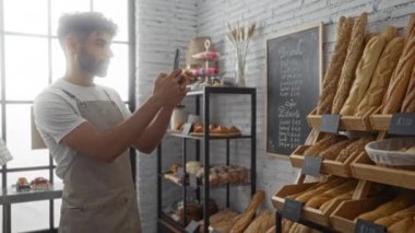 Sakallı genç adam fırında fotoğraf çekiyor. Taze ekmek ve hamur işleriyle dolu.