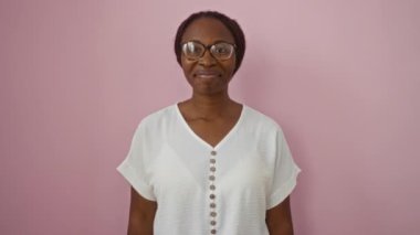 Genç Afrikalı Amerikalı kadın, gözlüklü, mutlu ve dostane bir gülümsemeyle pembe arka planda duruyor ve merhaba diyor.