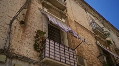 Mor çizgili tenteli eski bir balkon, saksı bitkileri ve berrak mavi gökyüzünün altında görünen pencere ve kablolarla barbar, puglia, İtalya 'da yıpranmış duvar..