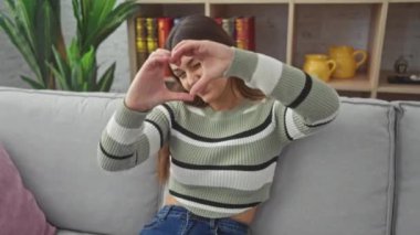 Yürek ısıtan ev sahnesi, çekici genç İspanyol kadın, kalp şeklinde el işareti ile sevgisini gösteriyor, sıcak oturma odasında ışıl ışıl gülümsüyor.