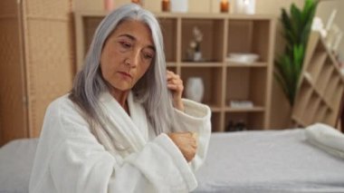 Samimi bir spa odasında saçları tarayan, beyaz bir bornoz giyen, gri saçlı, sakin ve rahat görünen bir sağlık merkezinde arka planda ahşap raflar ve bitkiler olan bir kadın.