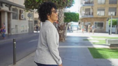 Genç, güzel, kıvırcık saçlı Afro-Amerikalı bir kadın gri bir ceketle şehir sokaklarında dikilirken kameraya gülümsüyor..