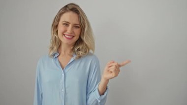 Tişört giyen neşeli, genç, sarışın bir kadın. Parmağını kenarı göstererek sevinçle gülümsüyor. Kameraya bakan el değmemiş, izole edilmiş beyaz bir arka planın önünde duruyor..