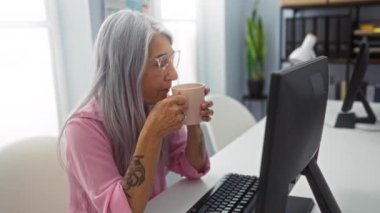 Ofiste bir fincan kahveyle çalışan bir kadın, gri saçlı, yetişkin, gözlüklü, pembe gömlekli, dövmeli, parlak, havadar bir çalışma odasında bilgisayar masasında oturan bir kadın.