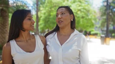 Anne ve kızı, yeşil yemyeşil ağaçlarla dolu bir parkta birlikte gülümsüyor. Bu da iki kadın arasında yakın bir aile bağı olduğunu gösteriyor.