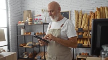 Ekmek ve hamur işleriyle çevrili fırının içinde tablet kullanan sakallı bir adam.