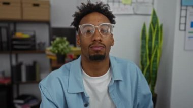 Modern ofis ortamında gözlüklü genç bir siyah adam görülüyor. Arka planda iç mekan bitkileri ve ofis malzemeleri var..