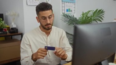 Çalışma alanı bilgisayar ve ofis bitkileri de dahil olmak üzere video görüşmesi sırasında elinde kredi kartı olan yakışıklı bir İspanyol..