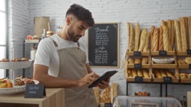 Fırında, raflarda ve tezgahlarda sergilenen ekmek ve hamur işleriyle çevrili bir tablet kullanan genç adam..