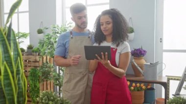 Önlüklü bir adam ve kadın güneşli bir çiçekçide bitkilerin arasında tablet kullanıyorlar.