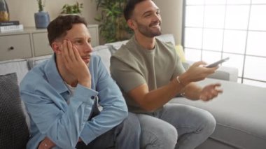 Oturma odasında elinde uzaktan kumandayla oturan iki adam ve diğeri sıkılmış görünüyor, rahat ve rahat bir ev ortamı sergiliyor.